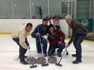 Three men and one woman smiling on ice rink with curling equipment