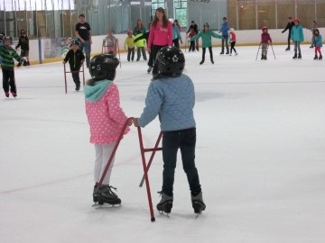 Two young girls ice skating with training aid in front of other skaters