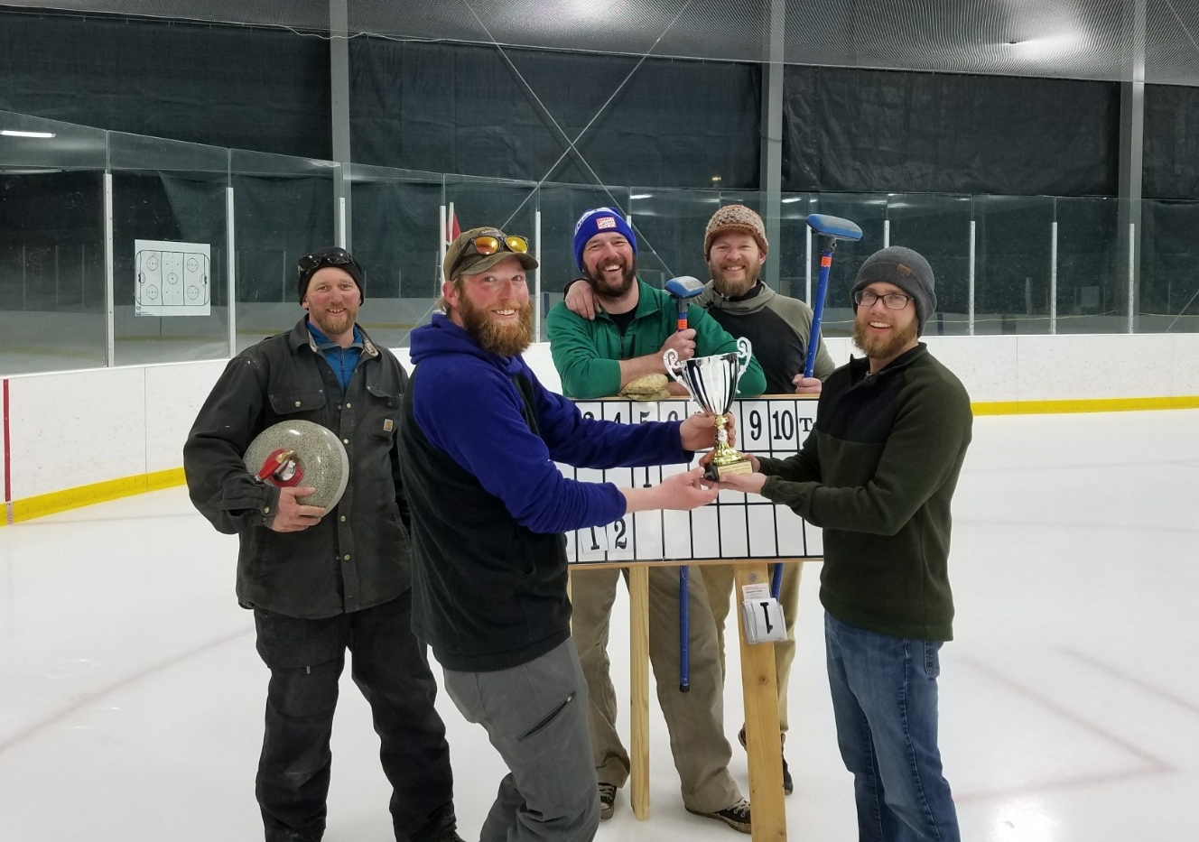 Five smiling males standing in ice rink with championship cup and curling equipment