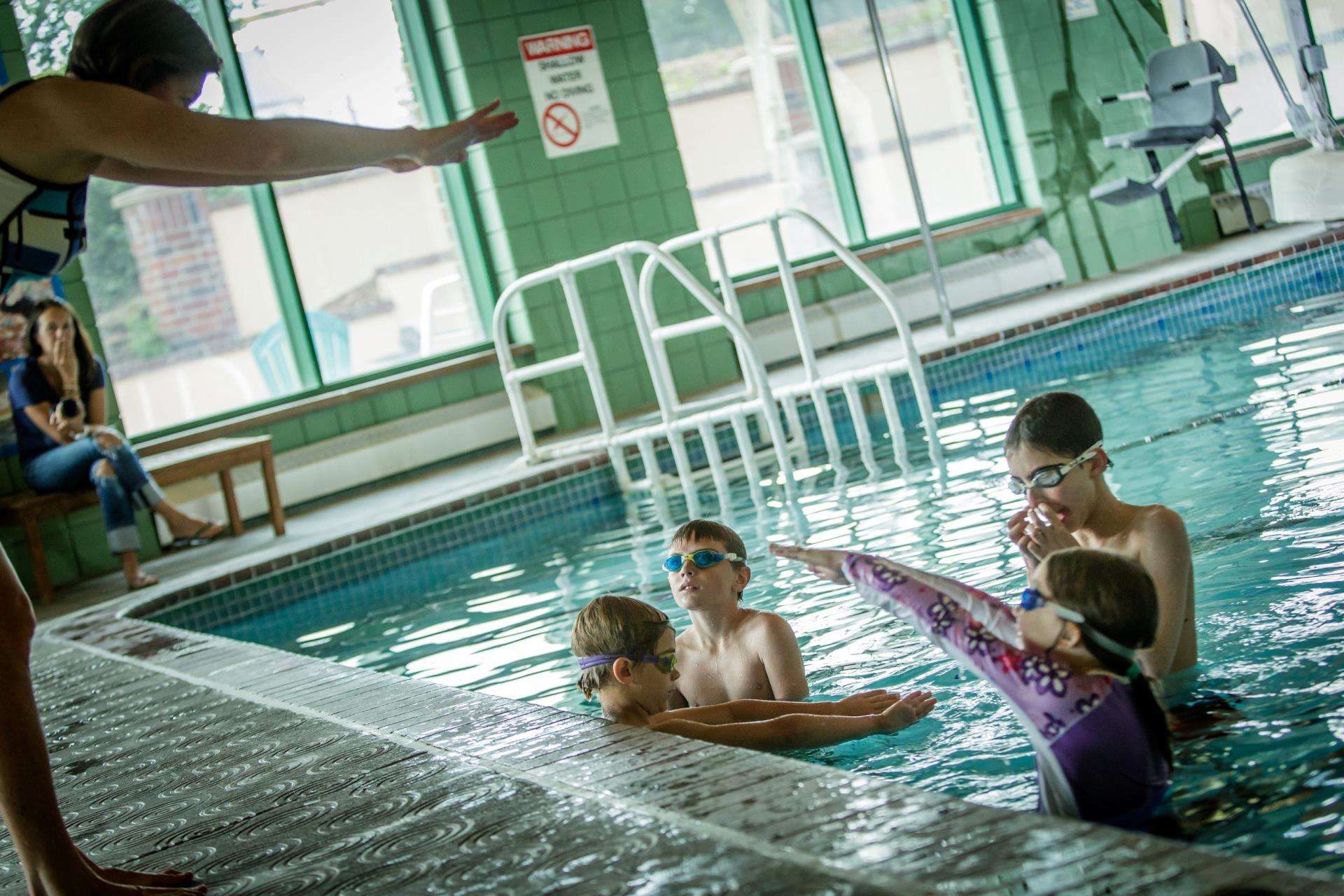 Group of 4 children in swim lesson in pool with instructor with diving arms on deck