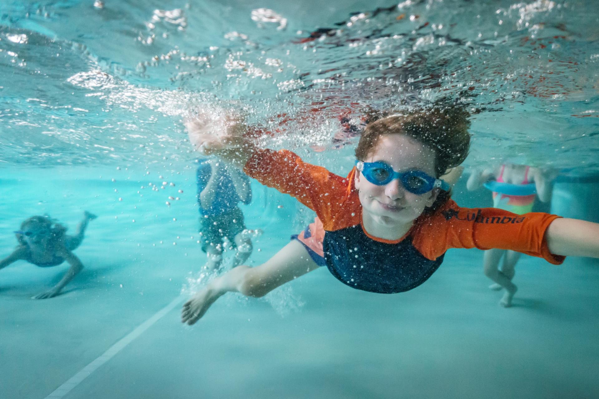 Boy in blue and orange shirt and goggles underwater with other children